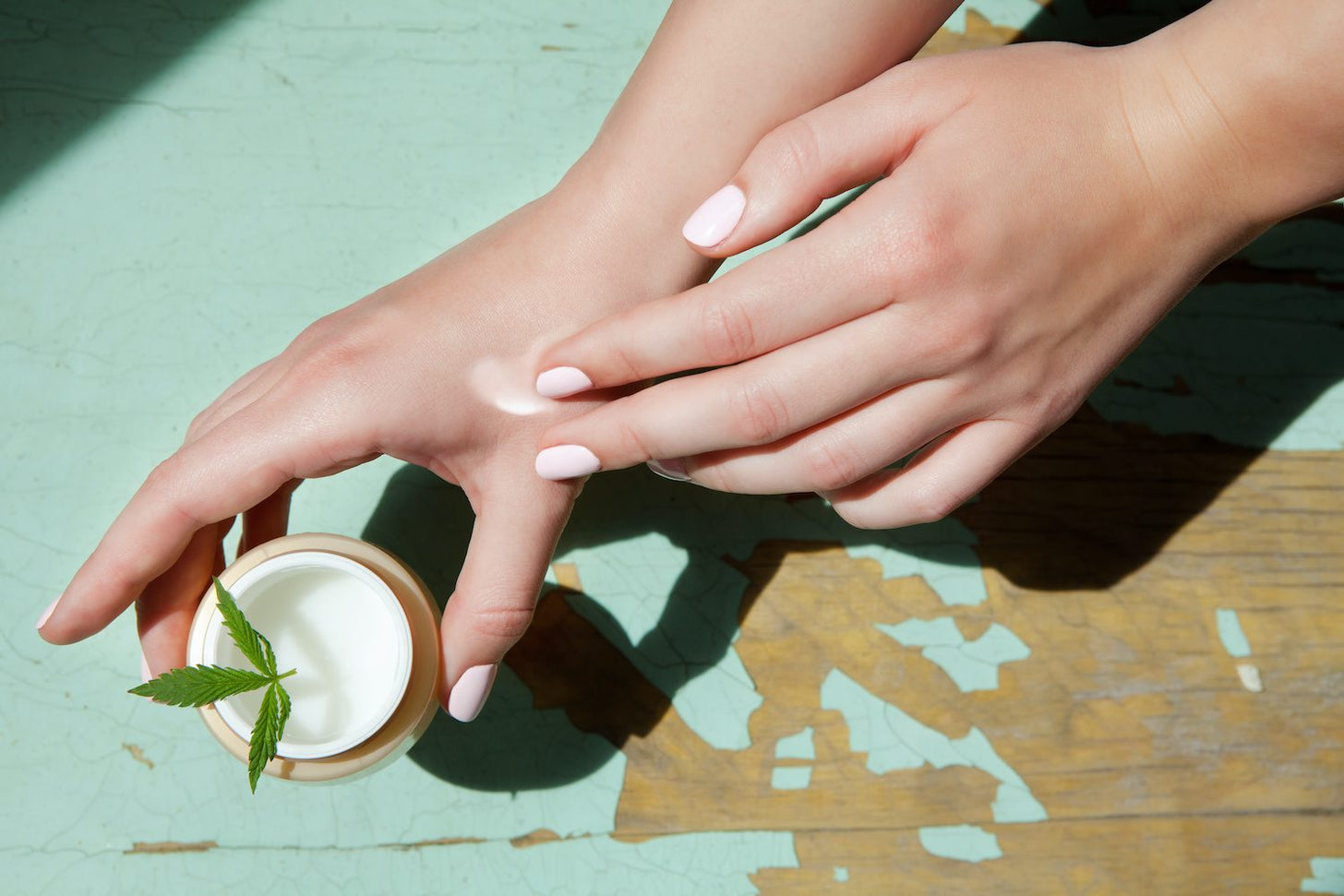 A woman holds a jar of cream in one hand while applying it to her skin with the other hand.