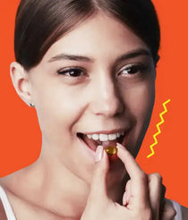 Young woman against an orange background bringing a yellow softgel supplement to her open mouth, smiling slightly.
