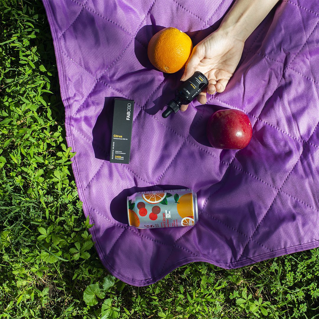 The corner of a purple blanket on green grass is shown with a seltzer, an apple and orange, and a person's hand holding a bottle of CBD oil in the dappled sunlight