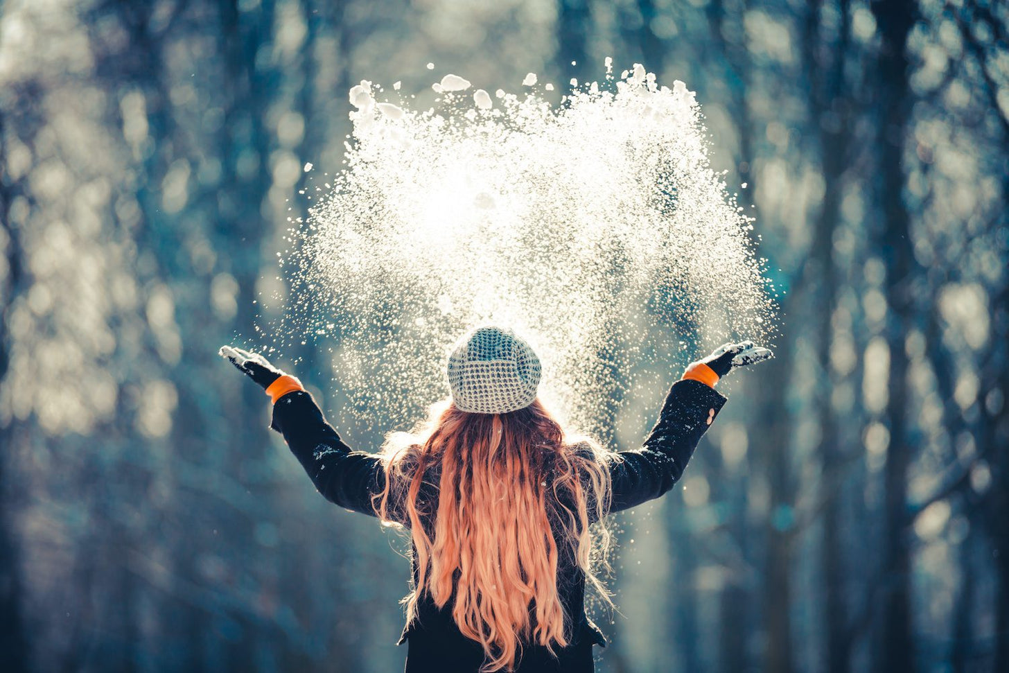 A woman with mittens an a hat stands outside looking at the trees and throws handfuls of powdery snow into the air