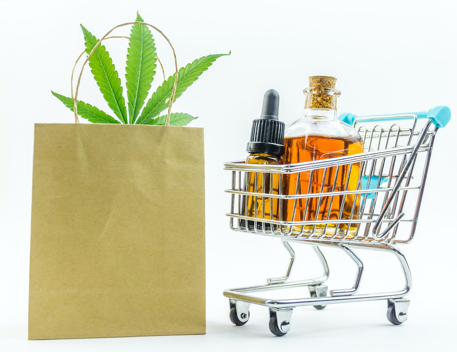 A tiny shopping cart with CBD oil bottles in it, next to a paper shopping bag with a hemp leaf poking out the top