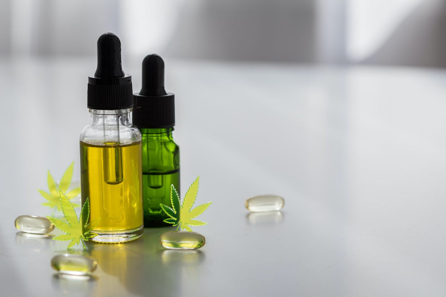 Two bottles of CBD oil stand on a white surface with gel capsules strewn around their bases