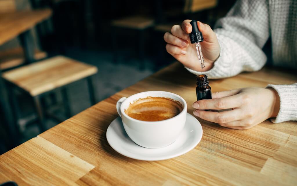 A latte in a white ceramic cup and saucer sits on a wooden table while a woman prepares a dropper of CBD to add to her coffee
