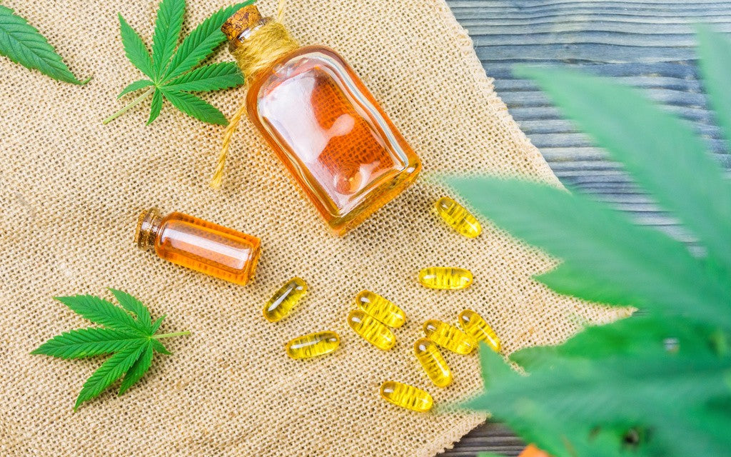 Hemp leaves, CBD capsules and bottles of CBD oil are scattered on a burlap cloth.