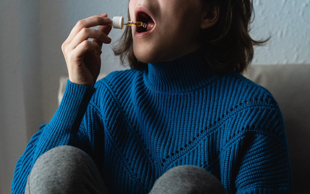A woman in a blue sweater sitting on a chair and taking a dropper of CBD under her tongue