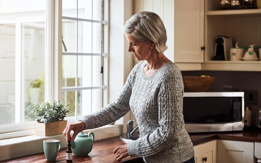 A woman stands at her kitchen counter and reaches for a bottle of CBD next to a teapot 