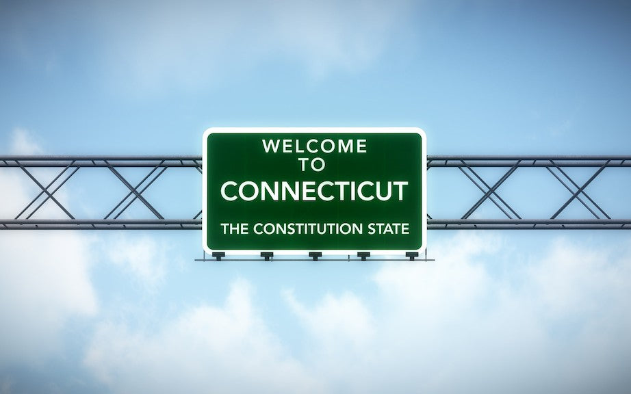 A highway sign with the blue sky behind it. The sign is deep green and says "Welcome to Connecticut. The Constitution State."