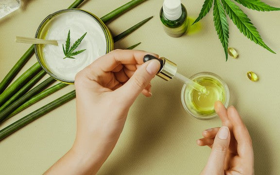 A woman holds a dropper of oil over a jar on a table, surrounded by soft gels, hemp leaves, and an open container of CBD cream