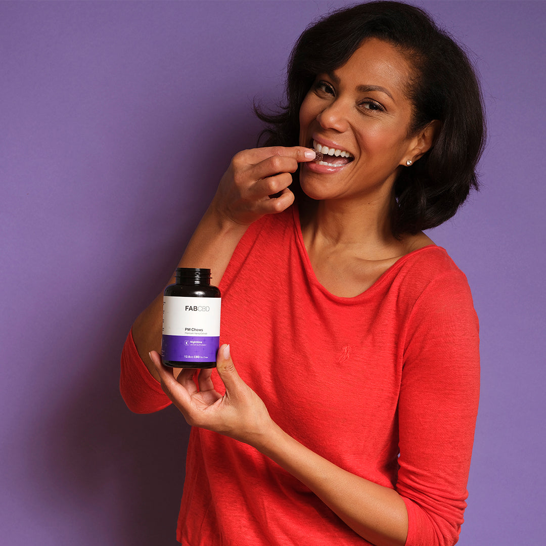 Woman in red shirt holding and about to eat a gummy from a purple and white Anytime + Sleep Gummy Bundle bottle.