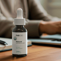 Amber glass dropper bottle of FAB CBD 'Natural' Broad Spectrum CBD Oil labeled '1200mg, 1 fl oz (30 mL), Dietary Supplement' with a green USDA Organic seal, placed on a wooden desk in front of a blurred person sitting at a desk with a notebook and pen, suggesting a calm workspace setting.