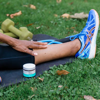 Close-up of a person stretching on a mat outdoors with a jar of Moisturizing Cream - 3 Pack visible on the grass.