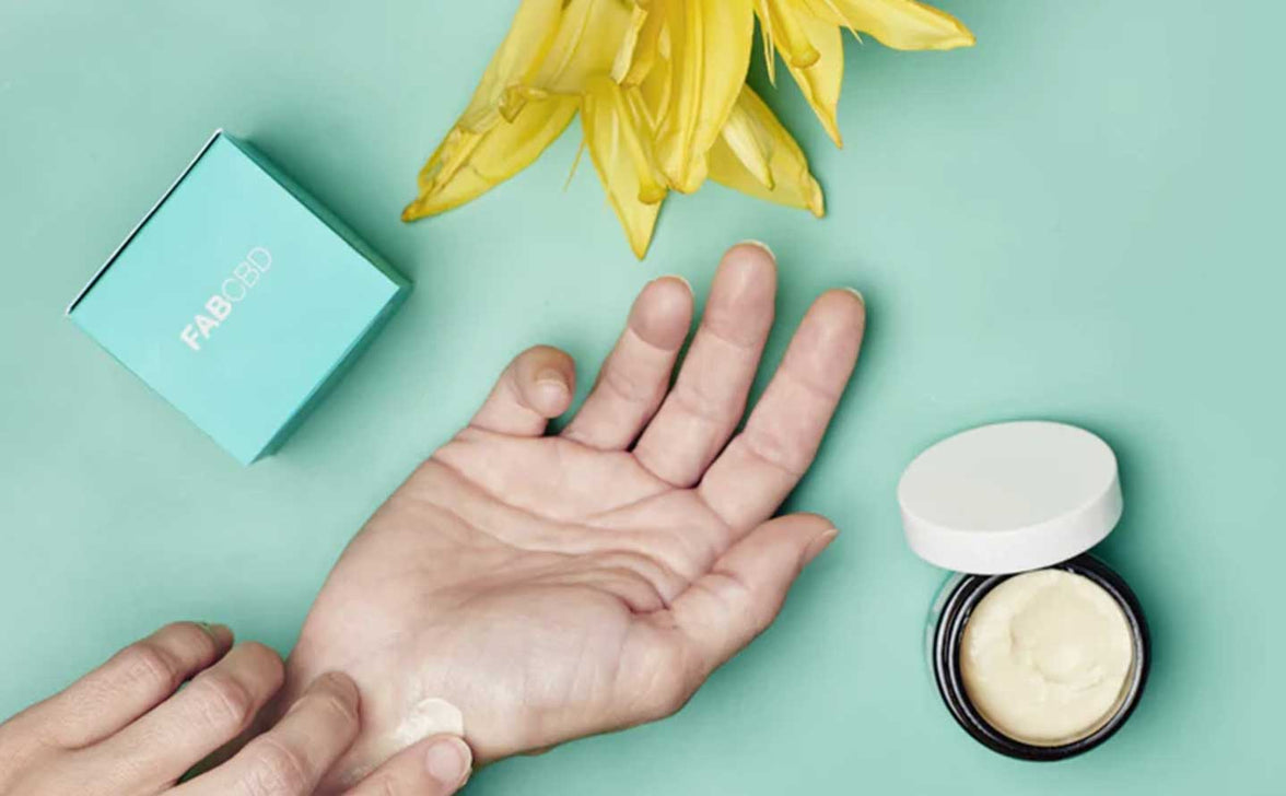 Top-down view of a hand applying Moisturizing Cream - 3 Pack beside an open jar and a turquoise FAB CBD box on a mint background.