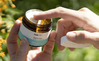 Close-up of hands dipping a fingertip into an open jar of Moisturizing Cream - 3 Pack outdoors, sunlight on the amber glass jar and green blurred background