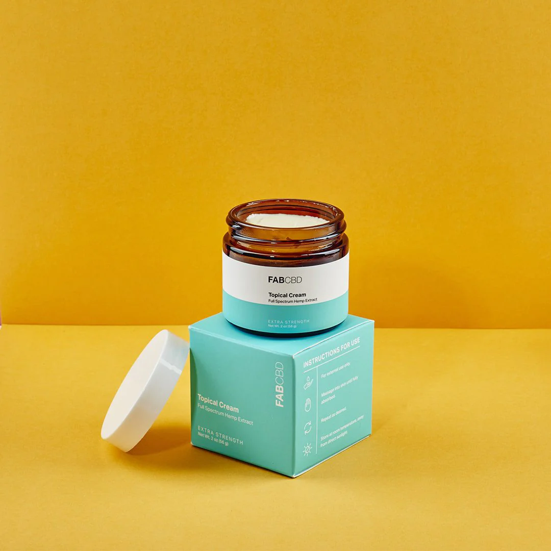 Center-front shot of an open FABCBD CBD Cream jar sitting atop its teal box with the white lid resting beside it against a warm yellow background.