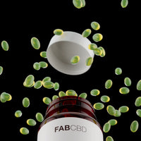 A close-up, dramatic view from below a FAB CBD Complete Cannabinoid™ 1:1:1:1 Softgels amber glass bottle, showing numerous translucent, dark green oval softgels floating and scattering in mid-air against a black background. The white cap of the bottle floats suspended above the opening, and the front of the bottle clearly displays the bold FAB CBD brand name.