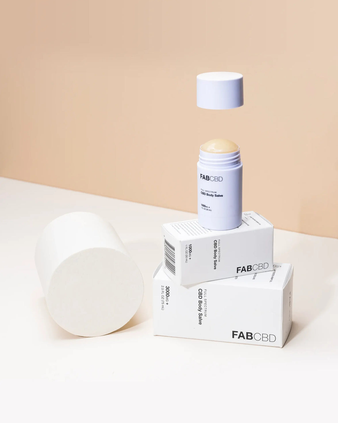 Close-up of a FABCBD CBD Body Salve stick with its cap hovering above the exposed balm, set on stacked white boxes against a beige backdrop.