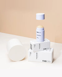 Close-up of a FABCBD CBD Body Salve stick with its cap hovering above the exposed balm, set on stacked white boxes against a beige backdrop.