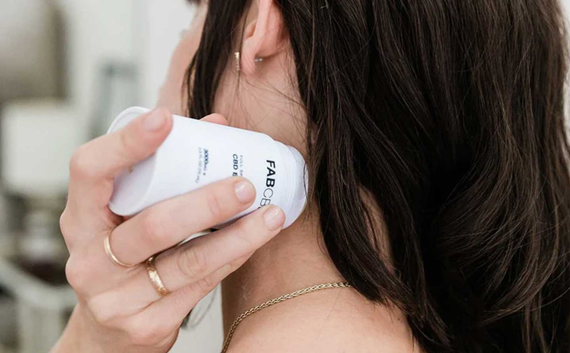 Close-up side view of a person applying the 2.5oz Salve - 3 Pack to their neck, the white jar held near the skin with soft natural light.