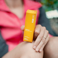 Close-up of a hand holding the FAB CBD CBD Oil citrus-orange box upright against a blurred pink jacket background.