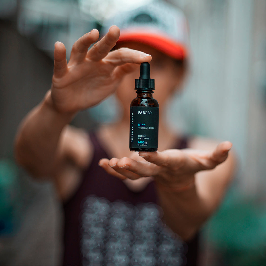 Centered close-up of a floating amber dropper bottle of CBD Oil held between a blurred person's palms and a shallow background.