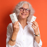 Smiling woman holds two white boxes of CBD Body Salve at chest level against an orange background, front-facing with soft even lighting