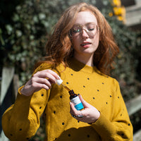 Close-up of a woman in a mustard polka-dot sweater holding a dropper and an amber bottle labeled "CBD Oil" at chest level.