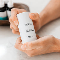 Close-up of hands holding a white FABCBD CBD Body Salve stick, shown at a slight angle against a soft, blurred background.
