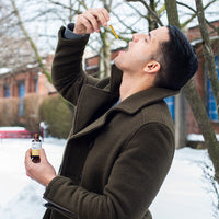 Side view of a man outdoors tilting his head back to drop CBD Oil into his mouth while holding the amber dropper bottle.