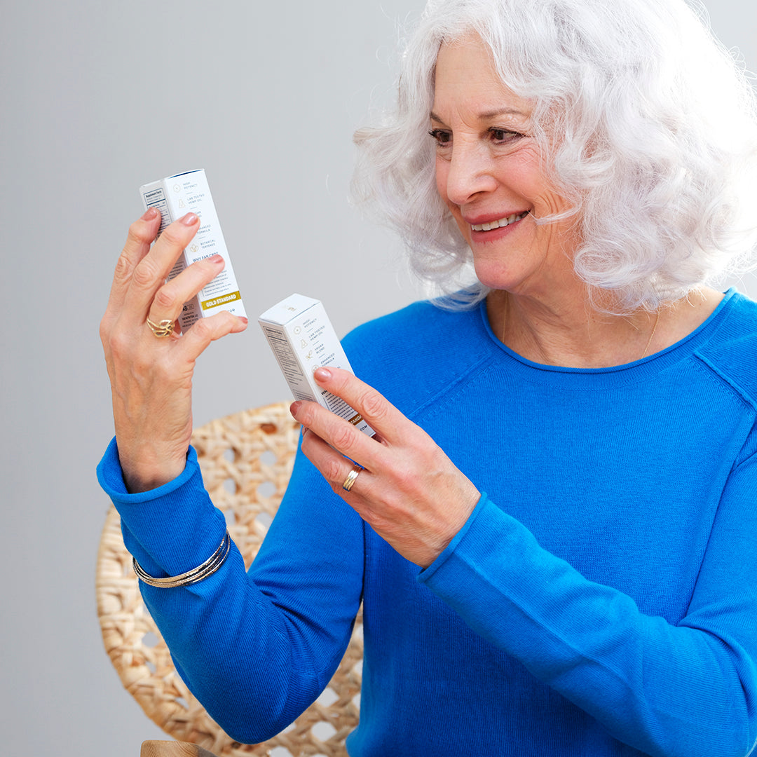 A smiling older woman with wavy white hair holding two white packaging boxes for FAB CBD 1:1 CBD+CBG Oil dietary supplement. Wearing a bright blue long-sleeved shirt, she looks closely at the box in her left hand, while her right hand holds the other box upright. The side panels of the boxes show consumers' product icons including high potency, lab tested hemp oil, vegan blend, and enhanced formula, with a gold "Gold Standard" band visible at the bottom against a soft gray background.