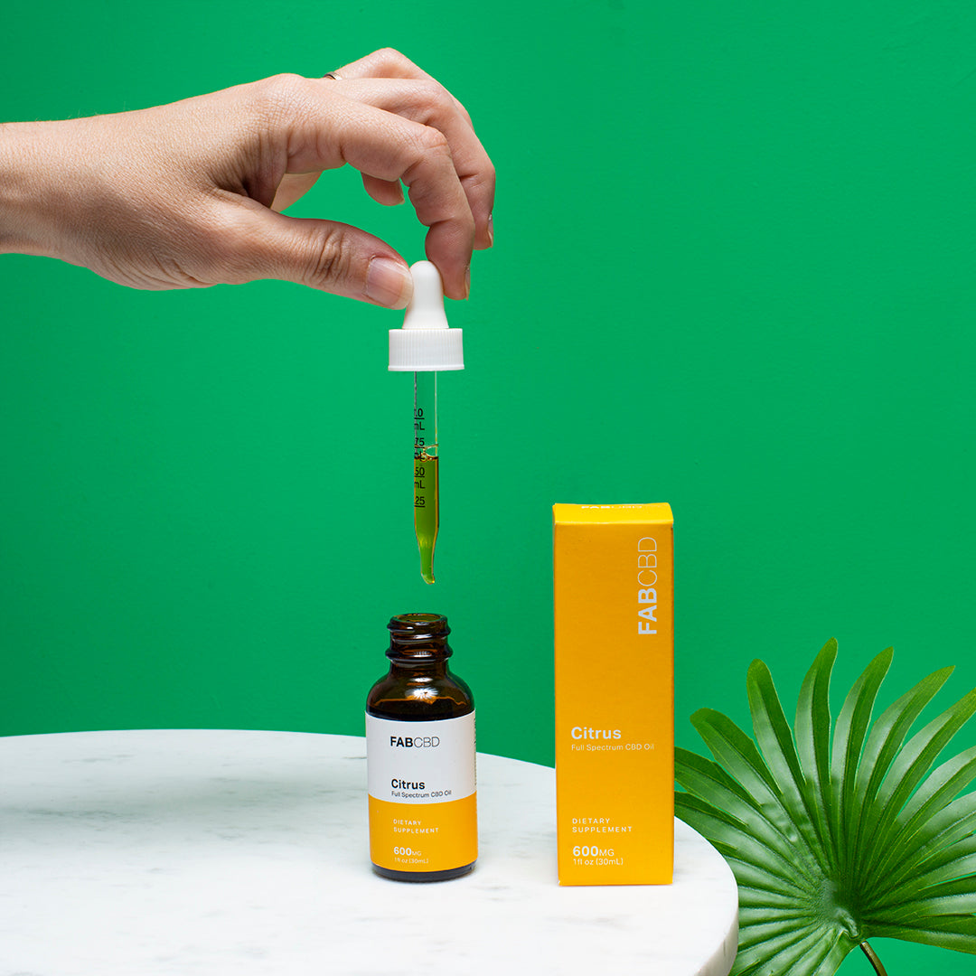 Top-down view of a hand holding the dropper over the FAB CBD CBD Oil Citrus bottle and matching yellow box against a bright green background.
