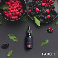 Top-down shot of a FABCBD CBD Oil dropper bottle centered on a dark matte surface surrounded by bowls of raspberries and blackberries and scattered mint leaves