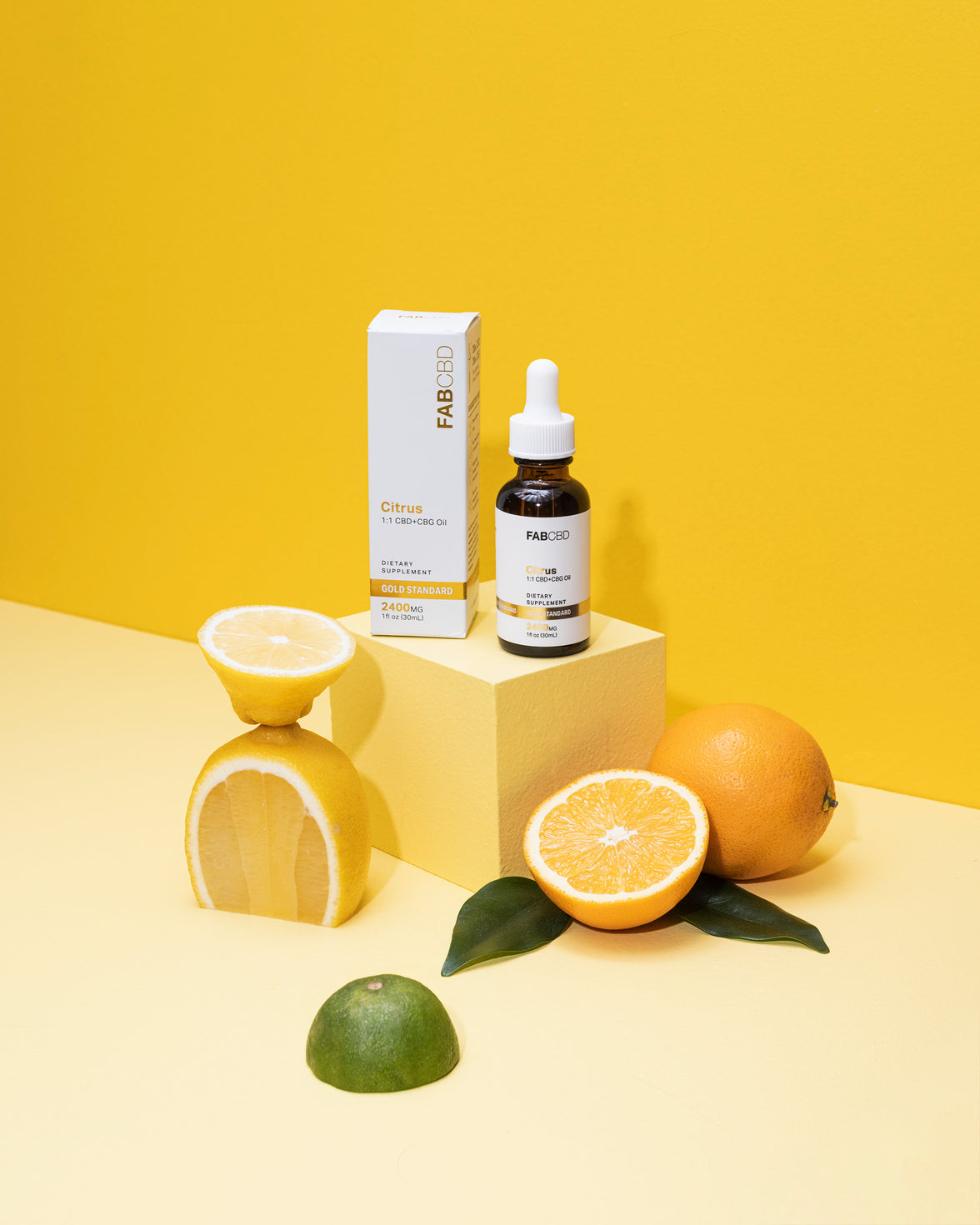 An amber dropper bottle and matching packaging box of FAB CBD Citrus 1:1 CBD+CBG Oil standing on a yellow display cube. The white labels feature a gold "Gold Standard" banner and specify "2400MG" and "1 fl oz (30mL)". Fresh citrus fruits—including a stacked cut lemon, a sliced orange with leaves, a whole orange, and a lime slice are arranged around the base against a bright yellow background.