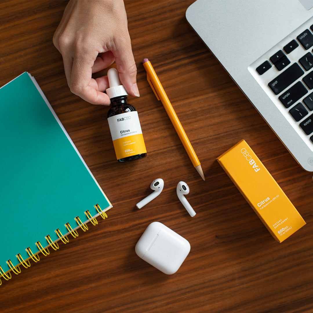 Top-down view of a hand picking up a FABCBD CBD Oil dropper bottle on a wood desk beside a yellow pencil, teal notebook, AirPods, and the product's orange box.