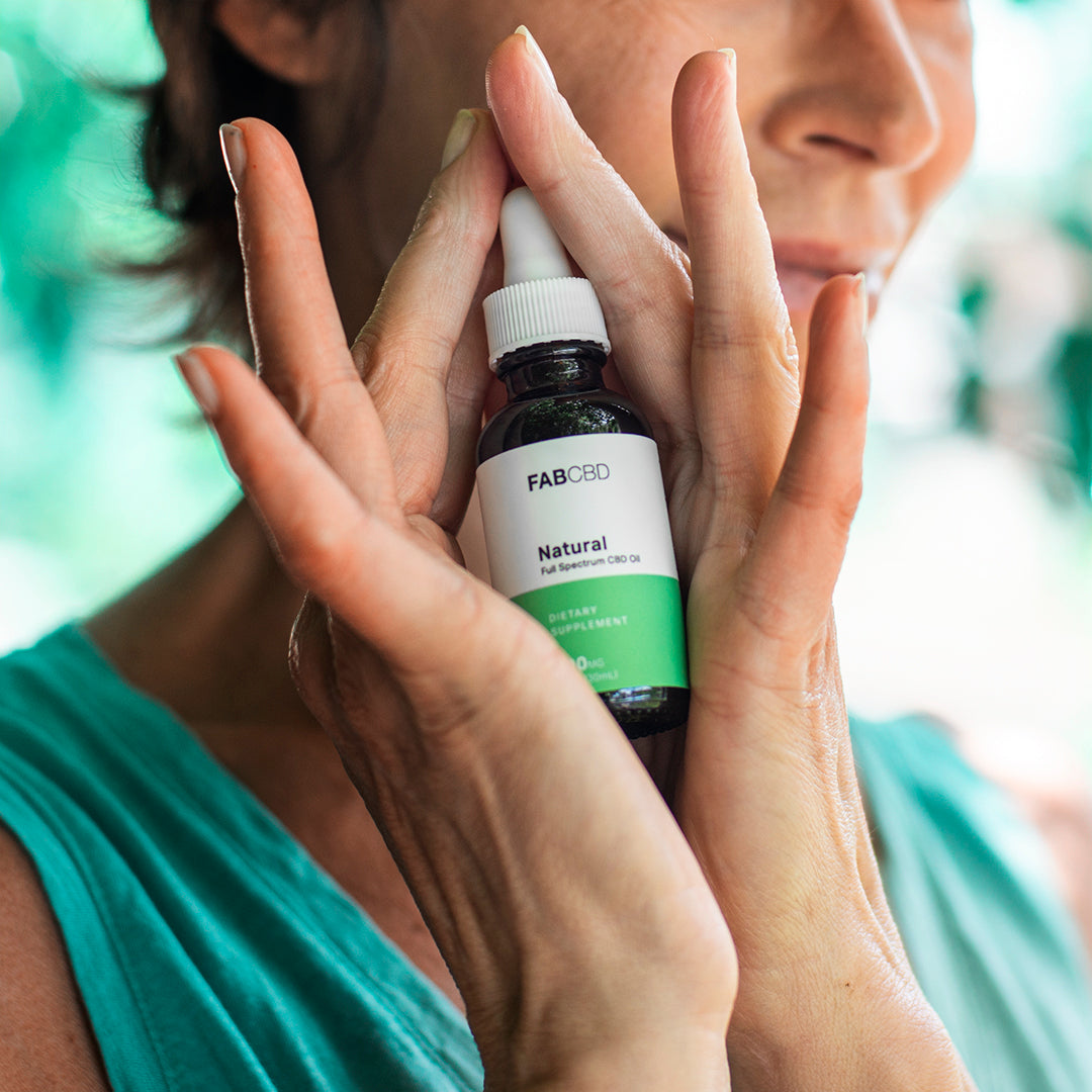 Close-up of a woman holding a bottle of CBD Oil by FABCBD between her palms near her face, soft natural light and a blurred green background.