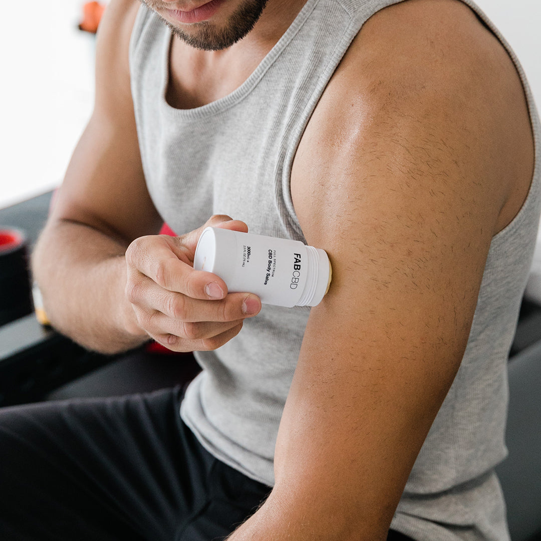 Close-up of a man applying FAB CBD CBD Body Salve to his upper arm from a white twist-up stick in soft, natural light.