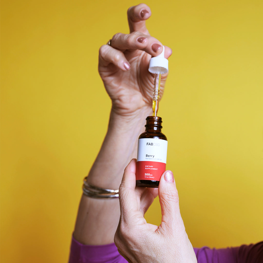 Close-up of a hand holding a CBD Oil dropper above its amber bottle against a bright yellow background.