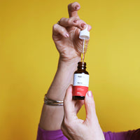 Close-up of a hand holding a CBD Oil dropper above its amber bottle against a bright yellow background.