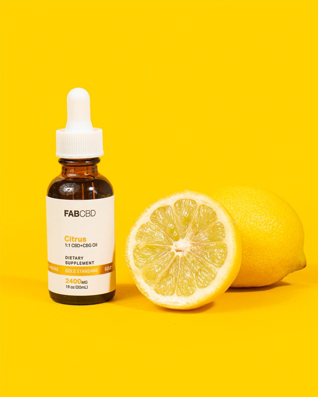 A vibrant product photograph featuring an amber dropper bottle of FAB CBD Citrus 1:1 CBD+CBG Oil dietary supplement. The white label includes a gold "Gold Standard" banner specifying "2400MG" and "1 fl oz (30mL)". Standing next to the bottle is a freshly halved juicy lemon, with a whole yellow lemon resting just behind it, all set against a matching solid bright yellow background.