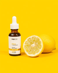 A vibrant product photograph featuring an amber dropper bottle of FAB CBD Citrus 1:1 CBD+CBG Oil dietary supplement. The white label includes a gold "Gold Standard" banner specifying "2400MG" and "1 fl oz (30mL)". Standing next to the bottle is a freshly halved juicy lemon, with a whole yellow lemon resting just behind it, all set against a matching solid bright yellow background.