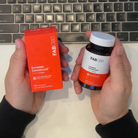 A top-down view of a person holding FAB CBD Complete Cannabinoid™ 1:1:1:1 Softgels products in their hands above a computer keyboard. The left hand holds the bright orange cardboard packaging box, while the right hand holds the amber glass supplement bottle with a white cap. Both the box and bottle labels display key product specifications: "CBC+CBG+CBD+CBN HEMP SUPPLEMENT," a potency of "15MG Per Serving," and a count of "60 PLANT BASED SOFTGELS."