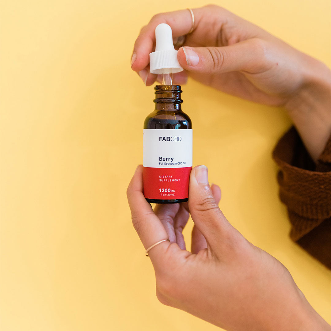 Close-up of hands holding a FAB CBD CBD Oil dropper bottle against a soft yellow background.
