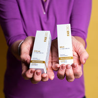 A person holding two white cardboard packaging boxes for FAB CBD 1:1 CBD+CBG Oil dietary supplements upright in their hands. The box on the left is labeled "Citrus" flavor and the box on the right is labeled "Mint" flavor. Both boxes display matching product specifications, including a gold "Gold Standard" band, "2400MG" potency, and "1 fl oz (30mL)" volume, set against a blurred purple shirt background.
