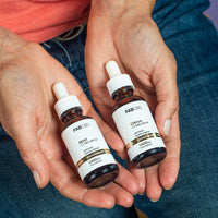 A person holding two amber dropper bottles of FAB CBD 1:1 CBD+CBG Oil dietary supplements cradled in their open palms. The bottle on the left is labeled "Mint" flavor and the bottle on the right is labeled "Citrus" flavor. Both white labels clearly show matching product specifications, including a gold "Gold Standard" band, "2400MG" potency, and "1 fl oz (30mL)" volume, set against a background of blue jeans.