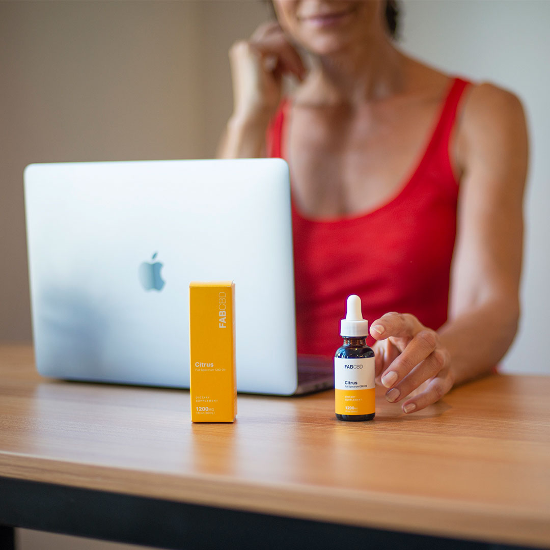 A shallow-focus shot of CBD Oil with a woman in a red tank top reaching for the amber dropper bottle beside its yellow box on a wooden desk.