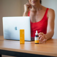 A shallow-focus shot of CBD Oil with a woman in a red tank top reaching for the amber dropper bottle beside its yellow box on a wooden desk.