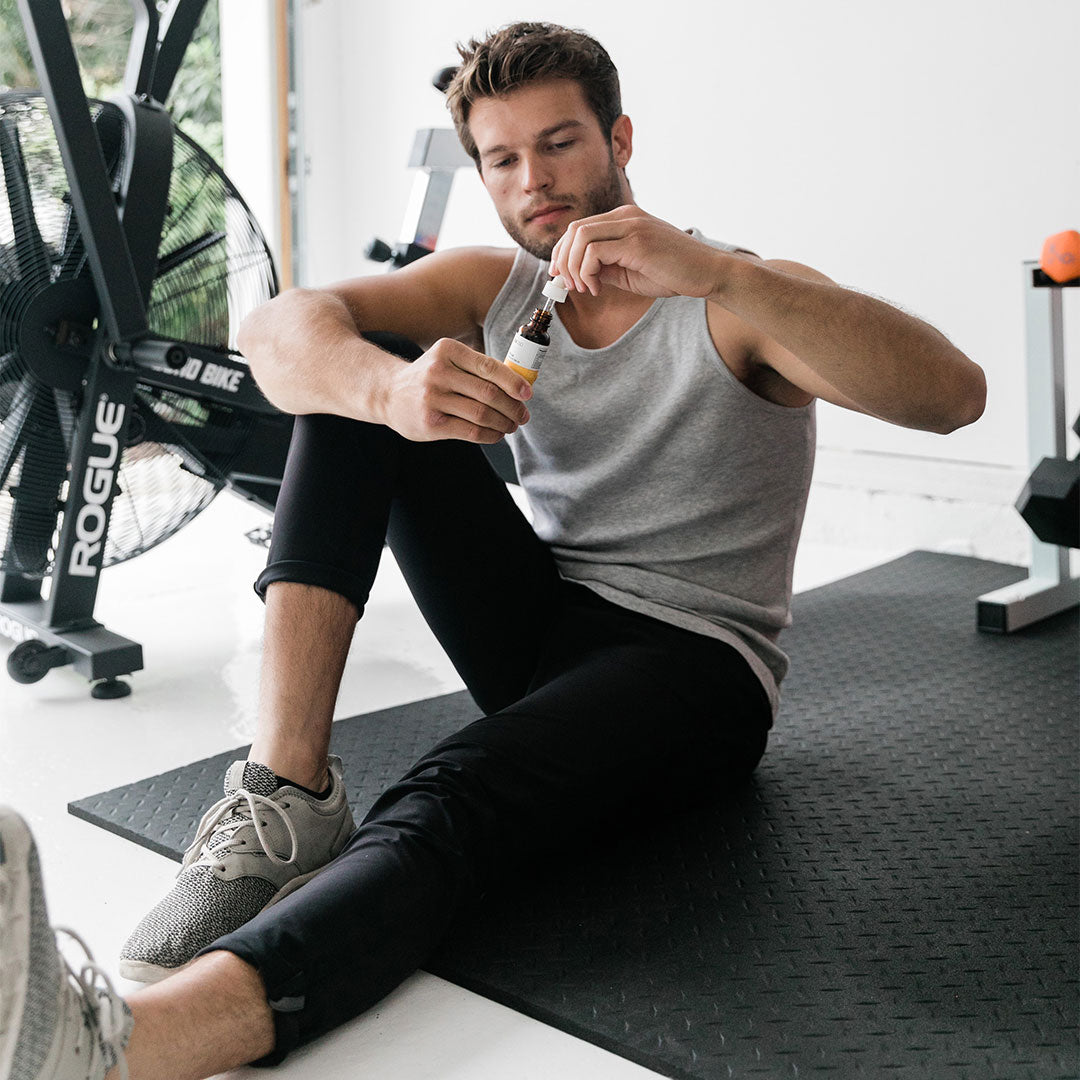 A seated man on a black gym mat holding a small amber dropper bottle of CBD Oil at eye level, shown in soft natural light with exercise equipment in the background.