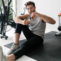 A seated man on a black gym mat holding a small amber dropper bottle of CBD Oil at eye level, shown in soft natural light with exercise equipment in the background.