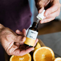 Close-up of a hand holding a FAB CBD CBD Oil dropper bottle above halved oranges, shot from a slight overhead angle.