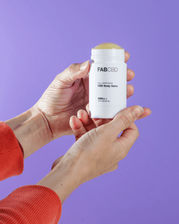 Hands holding a white stick of FAB CBD CBD Body Salve from a front angle against a lavender background, cap removed to show the balm.