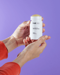 Hands holding a white stick of FAB CBD CBD Body Salve from a front angle against a lavender background, cap removed to show the balm.